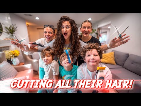 Six people in a bright living room pose with scissors and clippers, smiling for a playful hair-cutting moment.