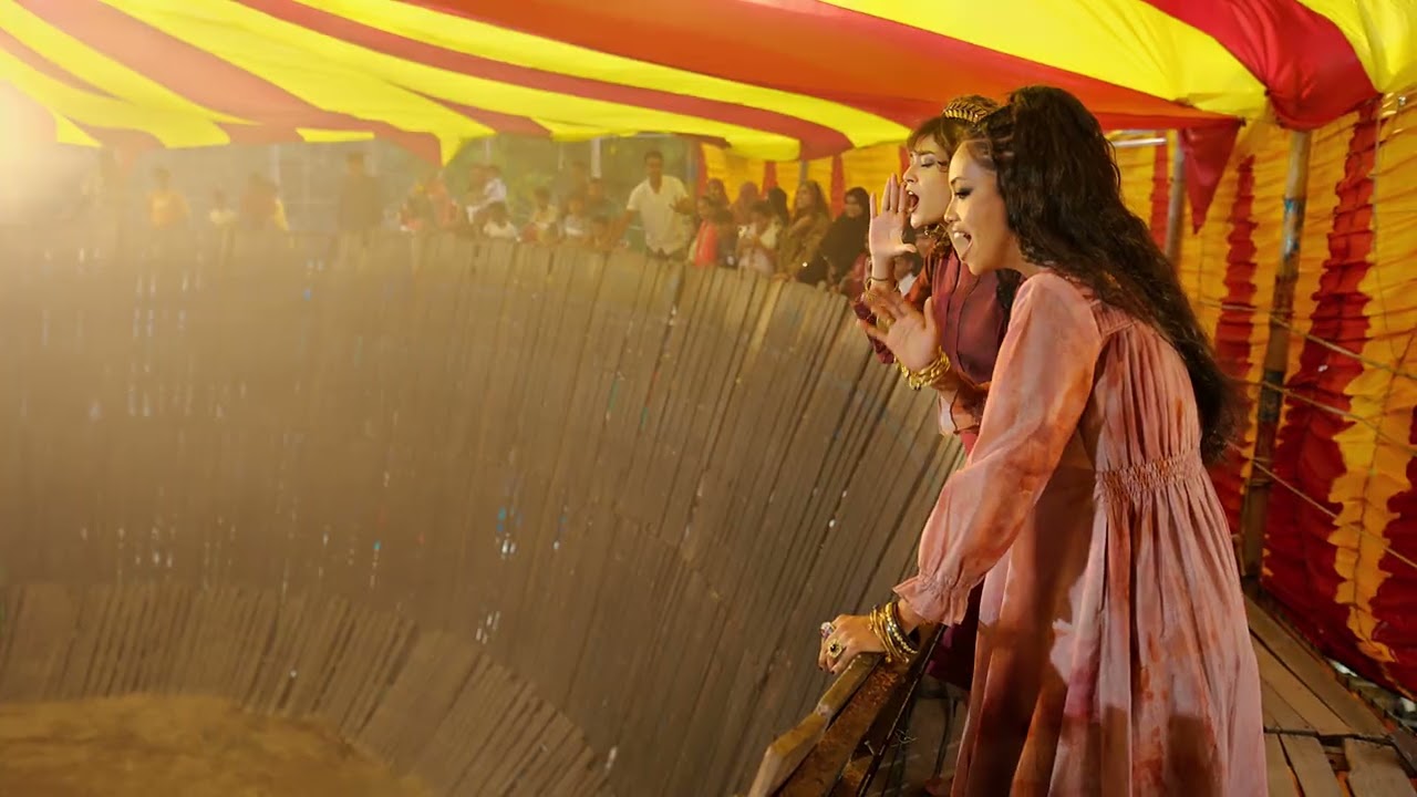 Four women in embroidered ethnic wear walk toward a brightly lit fairground stage at dusk