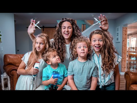 A woman with long curly hair holds up large scissors behind four children who are posing and smiling in a cozy living room, as if about to give a playful haircut.