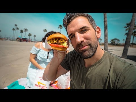 Smiling man on a sunny beach holds a burger toward the camera, with palm trees and a woman in a striped dress in the background.