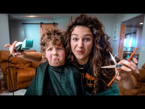 Woman cutting a child's hair at home, holding a pair of scissors and a blow dryer while the child sits in a cape in a cozy living room