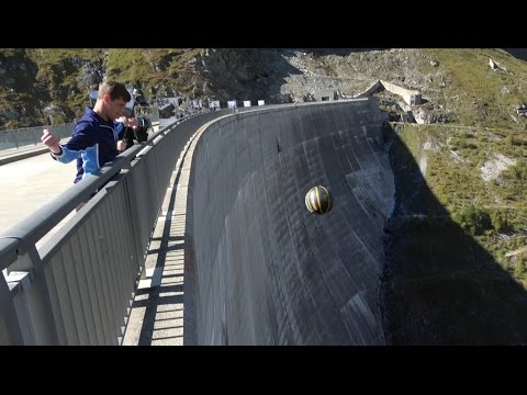 Young man stands on a bridge railing inspecting a black-and-yellow ball rolling down a tall concrete dam wall near a rocky mountainside.