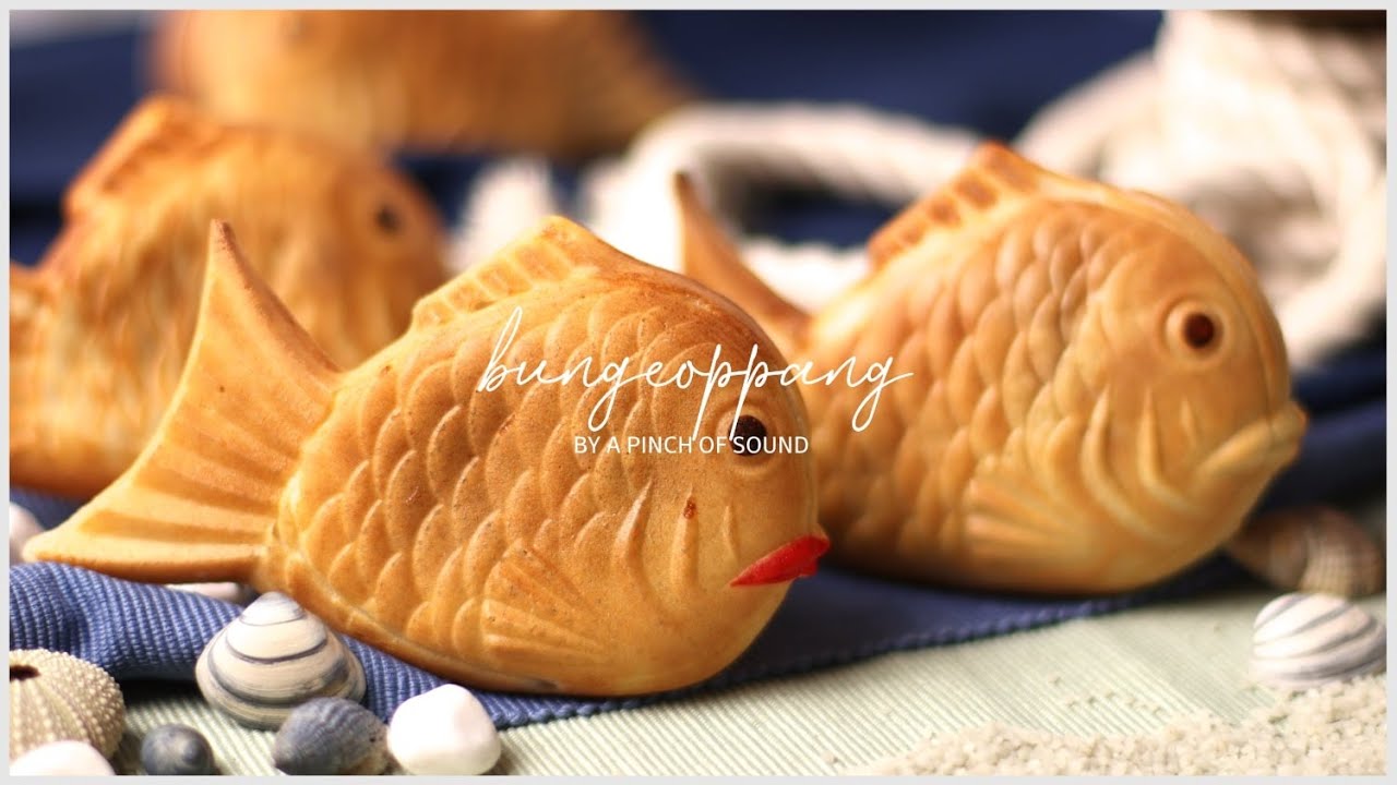 A close up of a hand holding a fresh hot Korean bungeoppang with red bean filling oozing out from the crispy edge of the fish-shaped bread