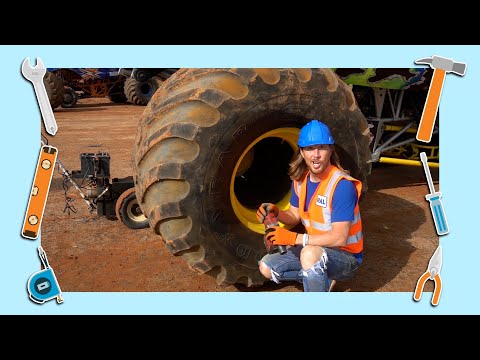 Mechanic in a blue hard hat and orange safety vest crouches beside a giant dusty tire, holding a power drill in a dusty yard workshop.