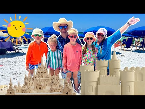Group of people on a sunny beach posing with intricate sandcastles and blue umbrellas in the background.