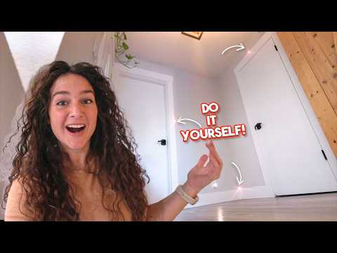 Woman with long curly hair smiling at the camera indoors, pointing to a bold 'Do It Yourself!' graphic on the wall.