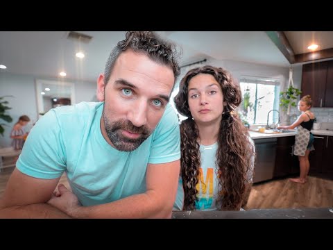 Couple leaning toward the camera in a bright modern kitchen, with a child in the background at a table and a woman at the sink nearby.