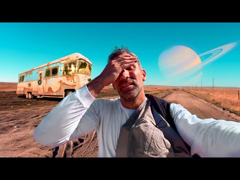 Selfie of a man on a dusty rural road, with an abandoned rusty bus to the left and a giant ringed planet in the blue sky behind him.