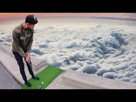 Man in a casual sweatshirt and dark pants stands on a small green practice mat on a rooftop, preparing a golf swing with clouds below at sunset.