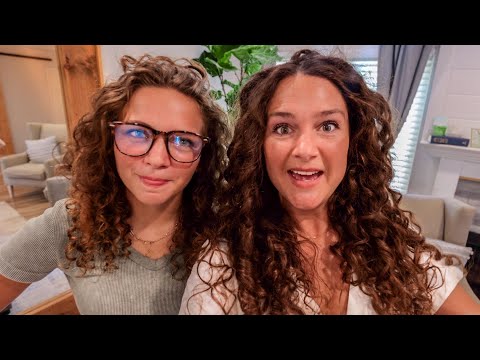 Two women with curly hair posing for a selfie in a cozy living room setting with lounge chairs in the background, smiling at the camera.