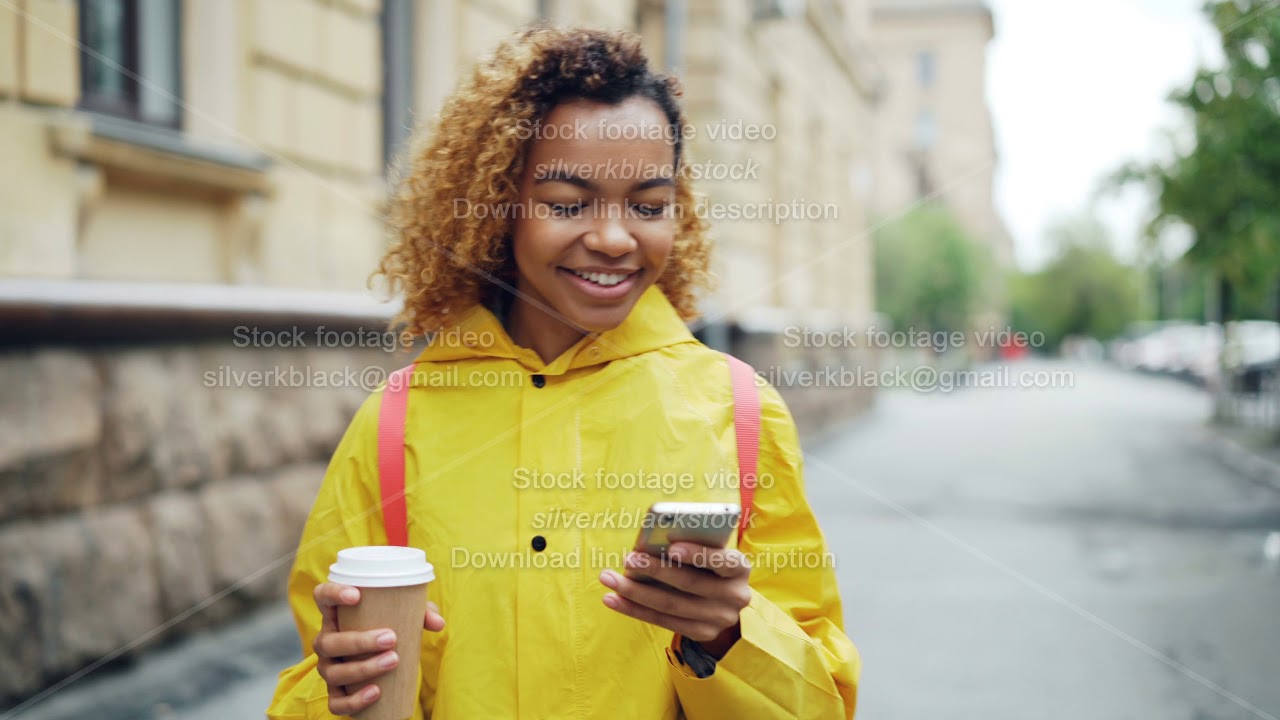 Happy African American woman is using smartphone touching screen and smiling walking outdoors in