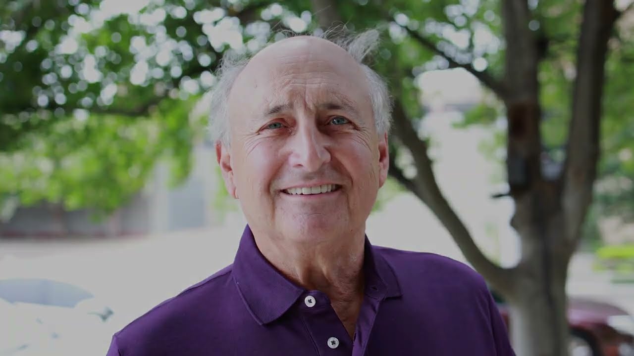 Senior man in purple polo shirt smiling with trees in background