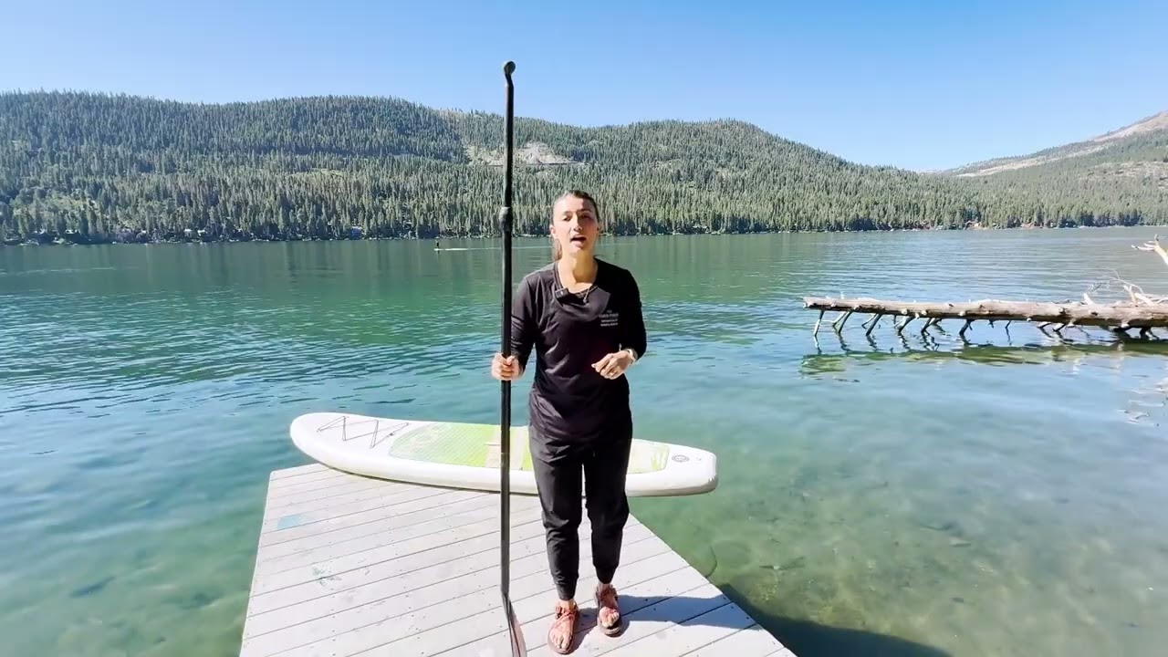 Woman standing on a dock at Donner Lake with a paddleboard on the dock and paddle in her hand.