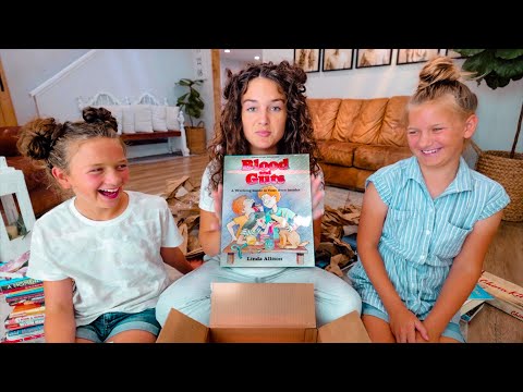 Three smiling girls sit on a living room floor, opening a box; the girl in the middle holds a book titled Blood Guts.