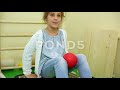 Little Girl Sits On Vibration Platform Squeezing Ball By Knees