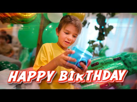 Young boy in a yellow shirt celebrates a birthday, holding a blue toy with green balloons and party decorations behind him; a 'HAPPY BIRTHDAY' banner is across the bottom.