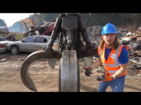 Construction worker in a blue hard hat and orange safety vest stands beside a giant hydraulic grapple in a scrapyard with wrecked cars in the background.