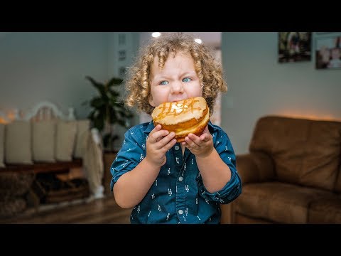 Curly-haired child in a blue shirt bites into a glazed doughnut indoors at home.
