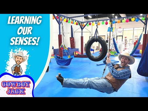 Cowboy-clad man swinging on a tire swing in an indoor play gym, with colorful equipment and a bold 'LEARNING OUR SENSES!' banner on the left dish side logo 'COWBOY JACK' visible.