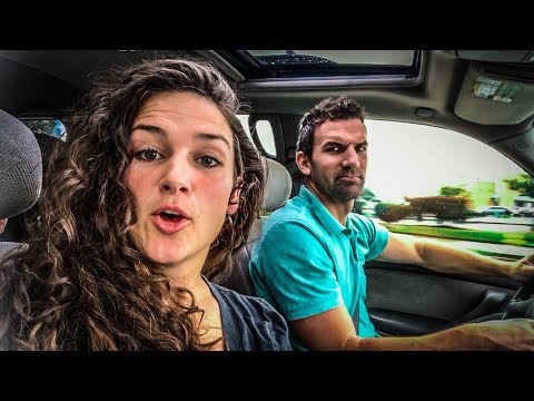 Two people in a car: a woman with curly hair in the passenger seat speaking to the camera while a man in a teal shirt drives.