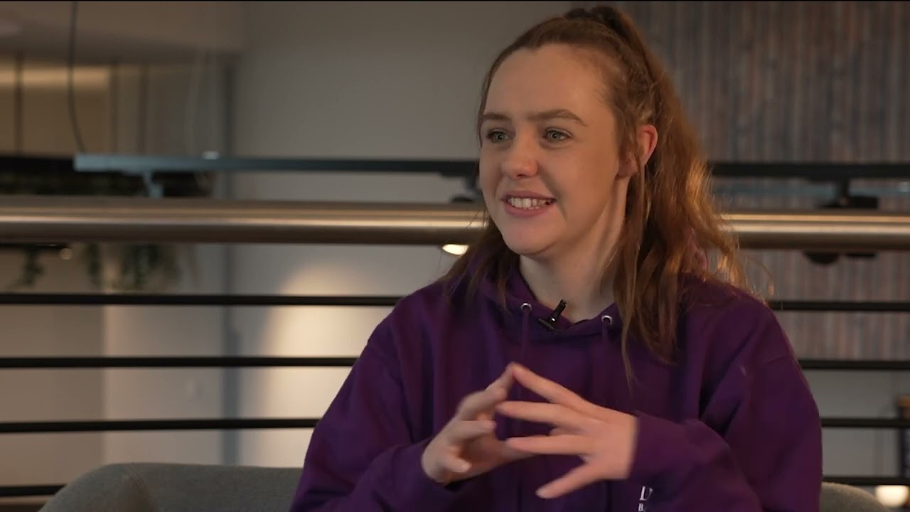 Student Rachel Price sits in our Basecamp venue, she is wearing a purple hoodie and has long brown hair tied up in a ponytail. 