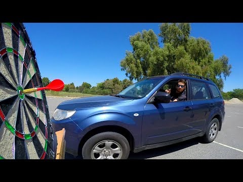 Man in a blue SUV aims a red dart at a large outdoor dartboard in a parking lot with a clear blue sky.
