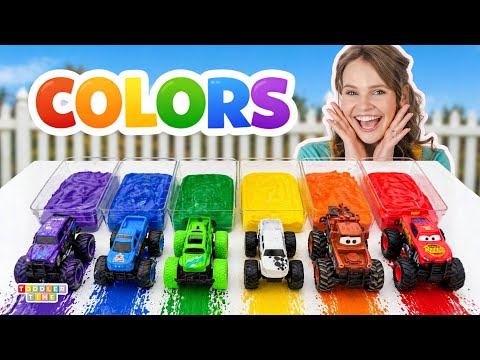 A cheerful girl sits beside rainbow-colored paint trays with six toy trucks in rainbow order, beneath a big 'COLORS' sign.