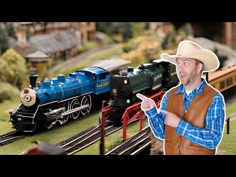 Man in a cowboy hat and brown vest points at blue and green model locomotives on a miniature railroad track set.