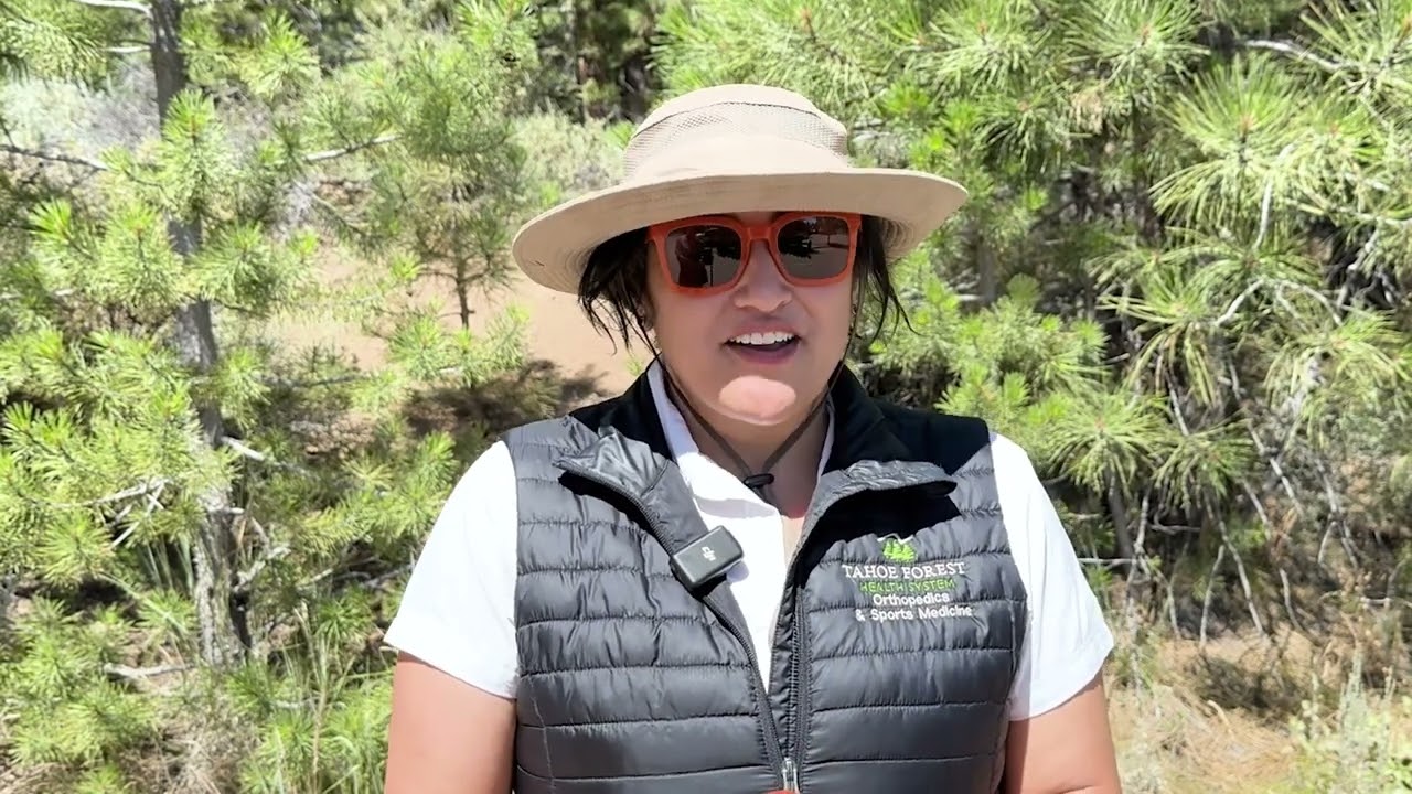 Woman wearing red sunglasses, a sun hat, white shirt and black vest on a forest trail