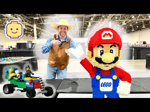 Smiling man in a cowboy hat and brown vest points at a giant LEGO Mario statue beside a colorful LEGO car at a convention hall.