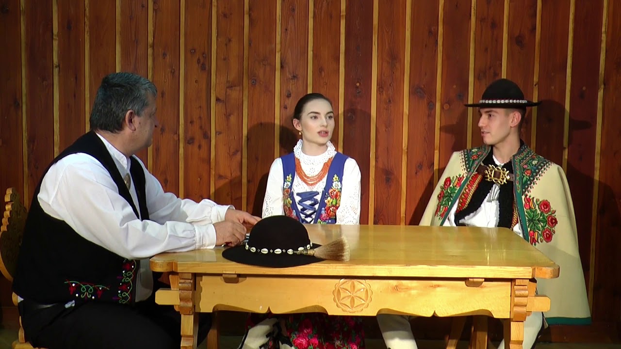 Dwóch mężczyzn i jedna kobieta w strojach podhalańskich siedzą przy stole, za nimi jest drewniana ściana