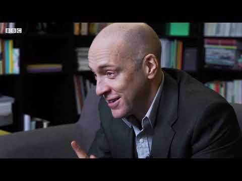 Derren Brown with talking in a room with a bookcase behind him.