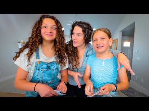 Three girls in a bright living room posing for a photo: one with curly hair in denim overalls, another with a gray hoodie, and the third in a blue striped shirt, all holding papers and smiling.