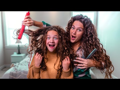 Two women with long curly hair having fun while styling hair; one holds a red curling iron and lifts the other's curls, both smiling in a bright bedroom.