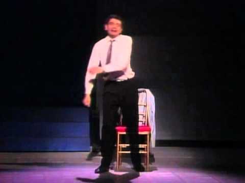 Rowan Atikinson in a shit, tie and black trousers on a stage.