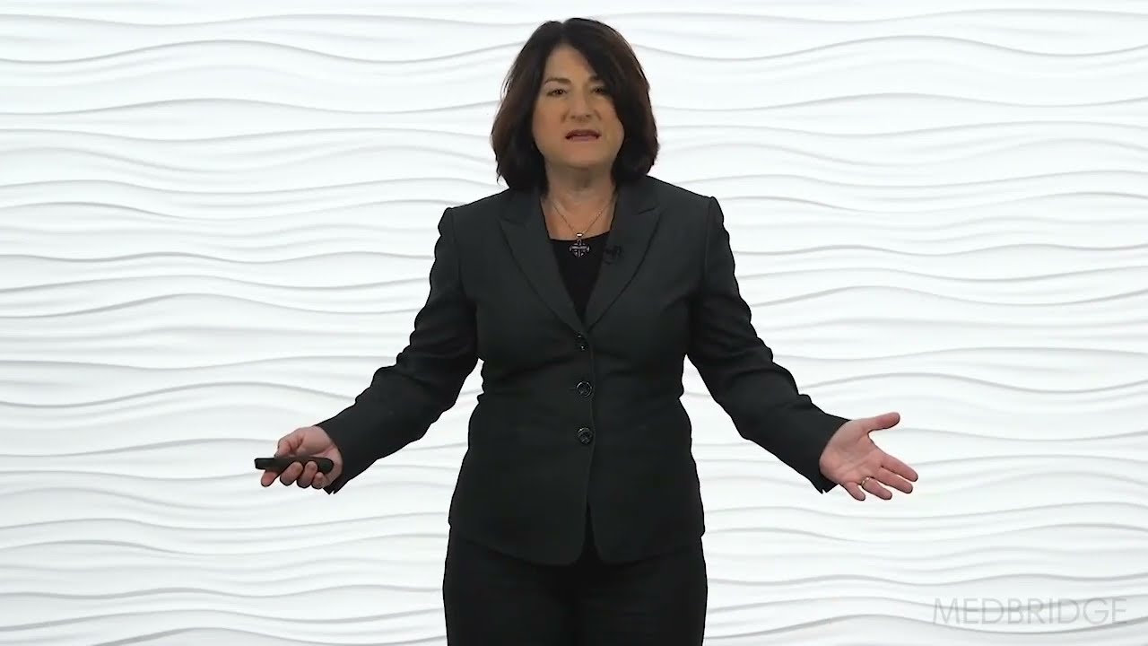 A person in a black suit stands against a wavy white backdrop, holding a clicker and gesturing to convey healthcare training.