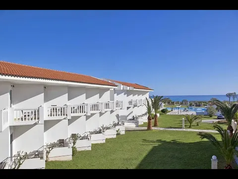 Außenansicht des Mrs Chryssana Beach Hotels mit weißen Gebäuden und roten Dächern, umgeben von grüner Rasenfläche und Palmen.