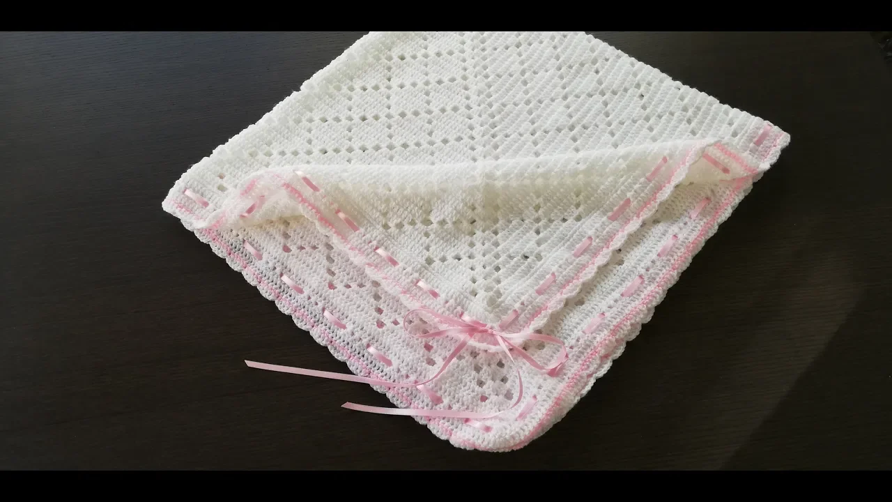 Manta de bebé a crochet blanca con rombos calados, borde rosado y cinta rosa, doblada sobre una mesa.