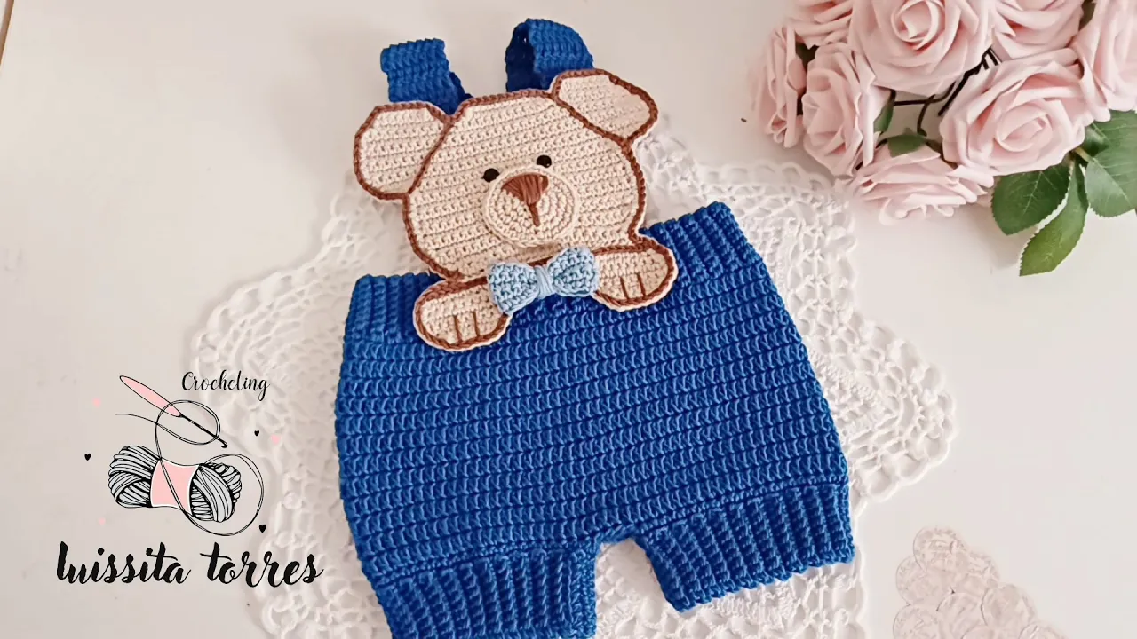 Jardinero de bebé azul a crochet con piernas cortas, tirantes y pechera de osito en beige.